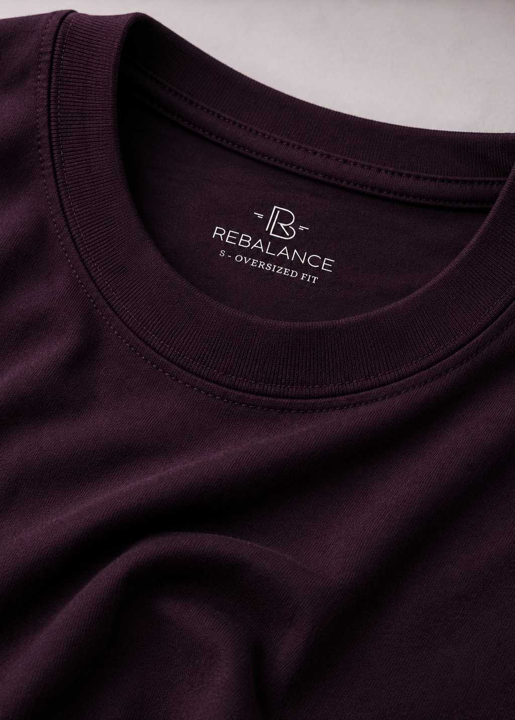 Close-up of a dark plum crewneck t-shirt showing the inner neck label with the white REBALANCE logo and '2 - OVERSIZED FIT' text.