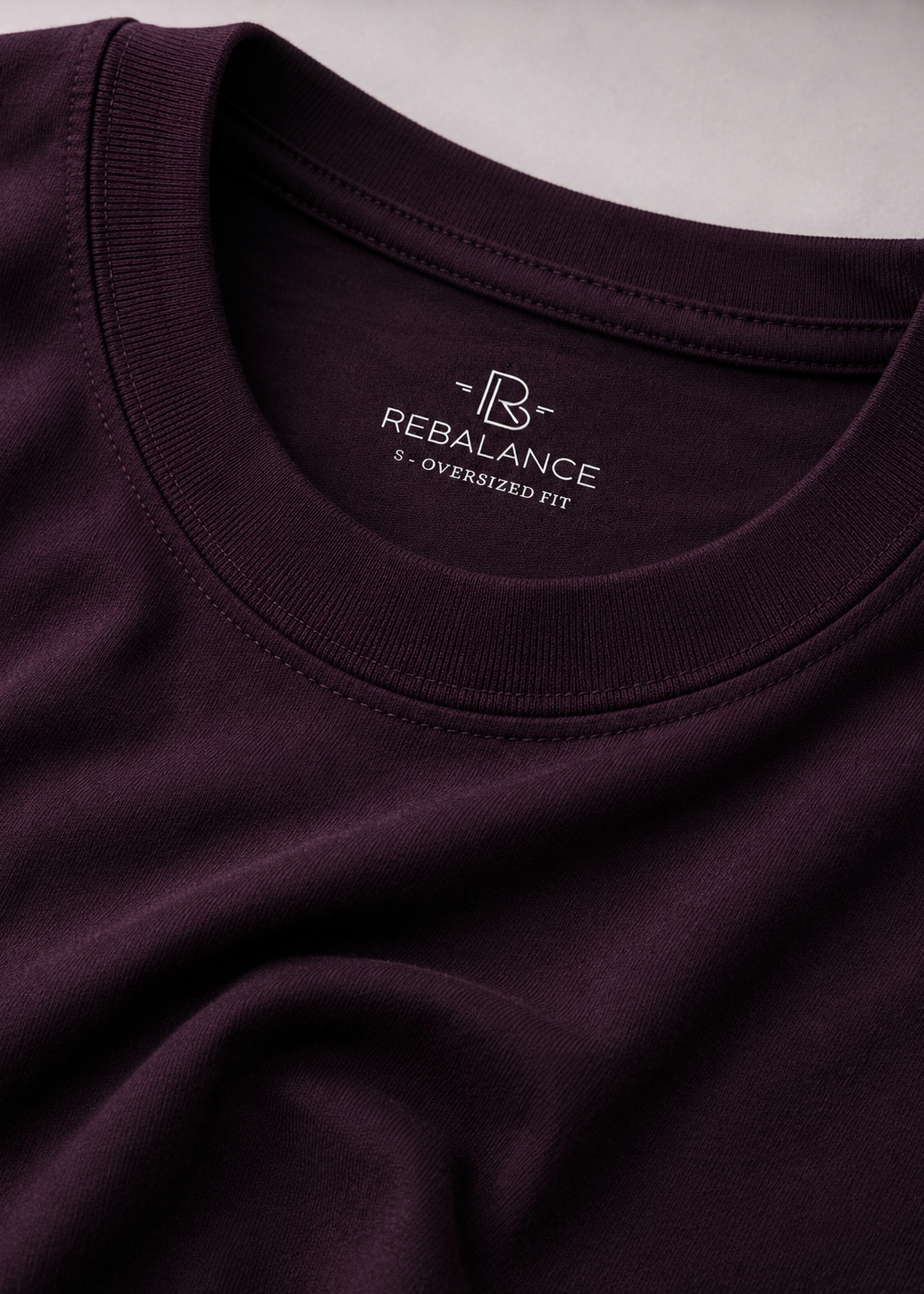 Close-up of a dark plum crewneck t-shirt showing the inner neck label with the white REBALANCE logo and '2 - OVERSIZED FIT' text.