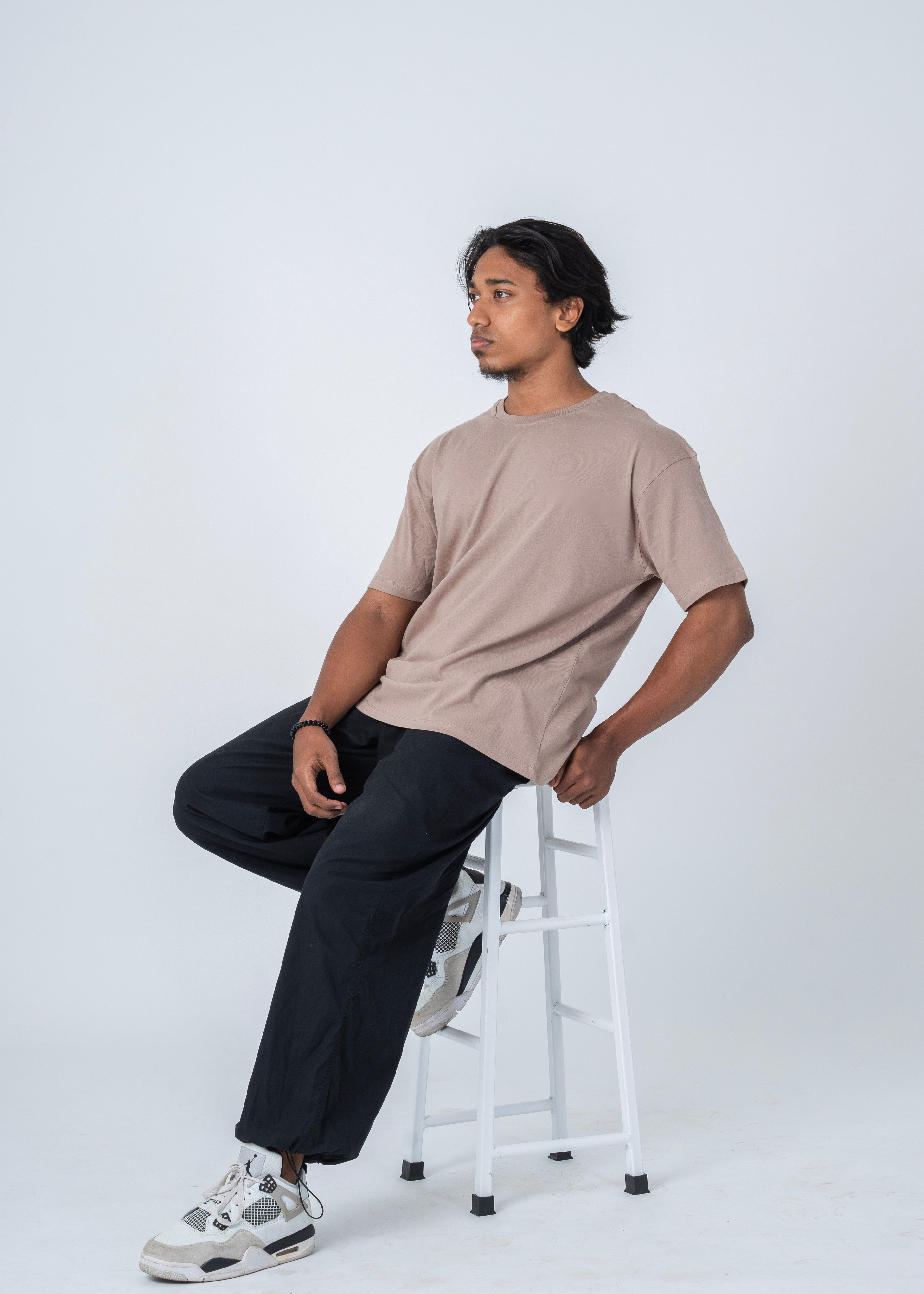 A man wearing a light beige crew-neck t-shirt, dark charcoal pants, and white sneakers sits on a white metal stool against a light gray studio background.
