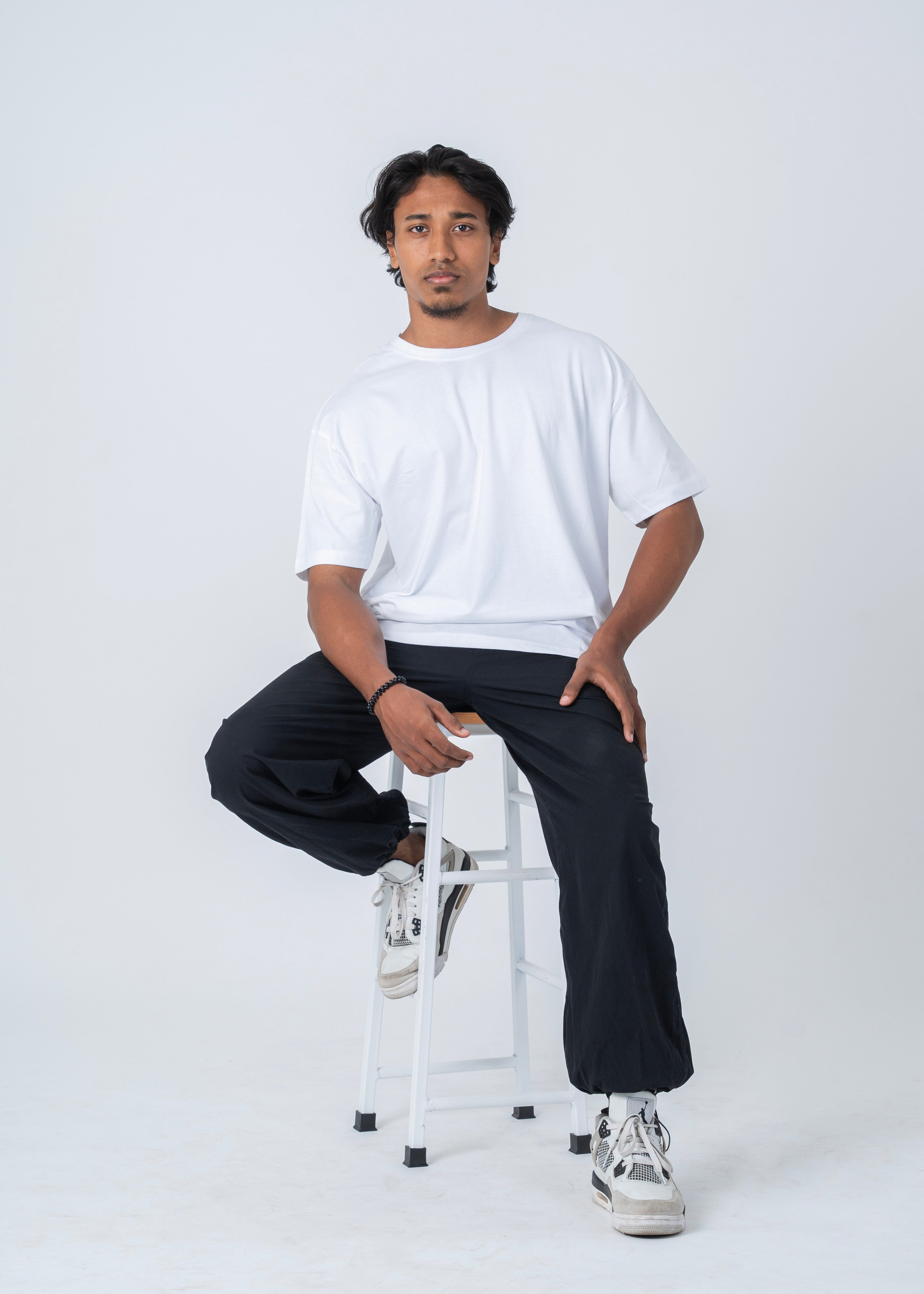 A man wearing a white crewneck t-shirt, black wide-leg cargo pants, and white sneakers, sitting on a white metal stool against a light gray studio background.