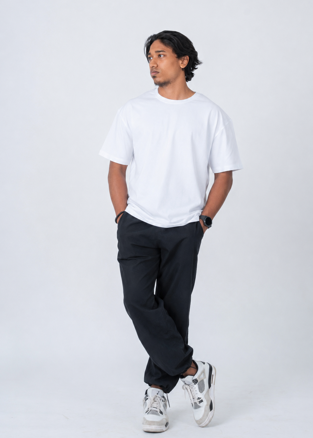 A man wearing a plain white short-sleeve t-shirt, loose black pants, and white sneakers, standing against a light gray studio background.