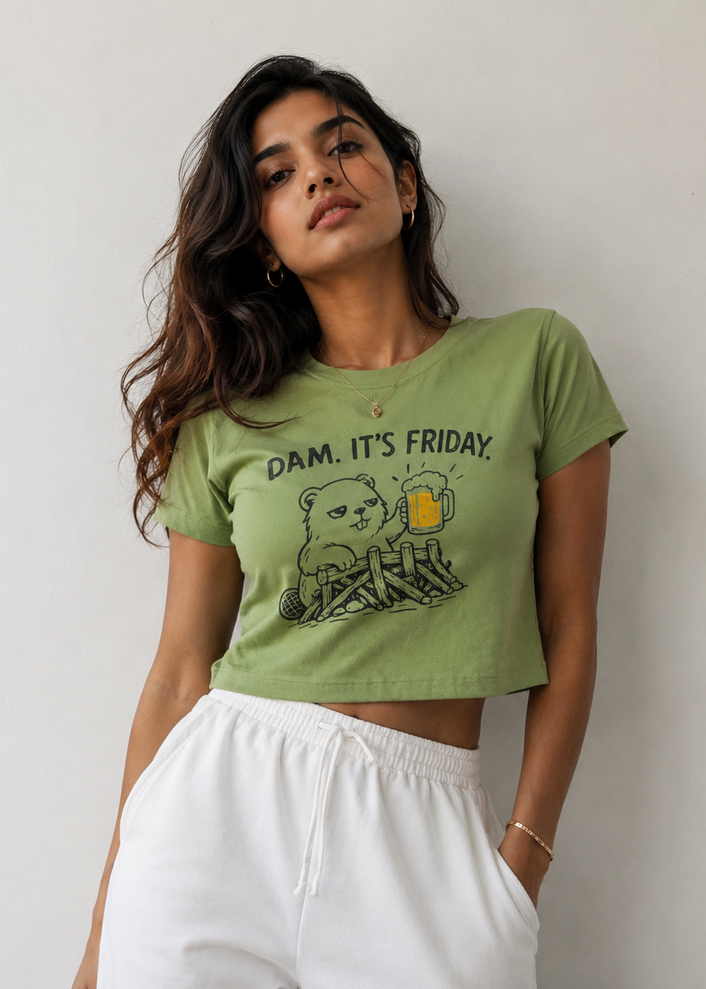 A woman wearing a light olive green cropped t-shirt with a cartoon beaver graphic and the text 'DAM. IT'S FRIDAY.
