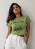 A woman wearing a light olive green cropped t-shirt with a cartoon beaver graphic and the text 'DAM. IT'S FRIDAY.