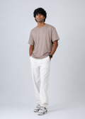A man wearing a taupe short-sleeve crewneck t-shirt, white trousers, and gray sneakers, standing against a light gray studio background.