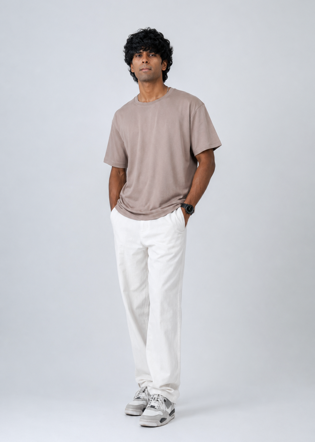 A man wearing a taupe short-sleeve crewneck t-shirt, white trousers, and gray sneakers, standing against a light gray studio background.