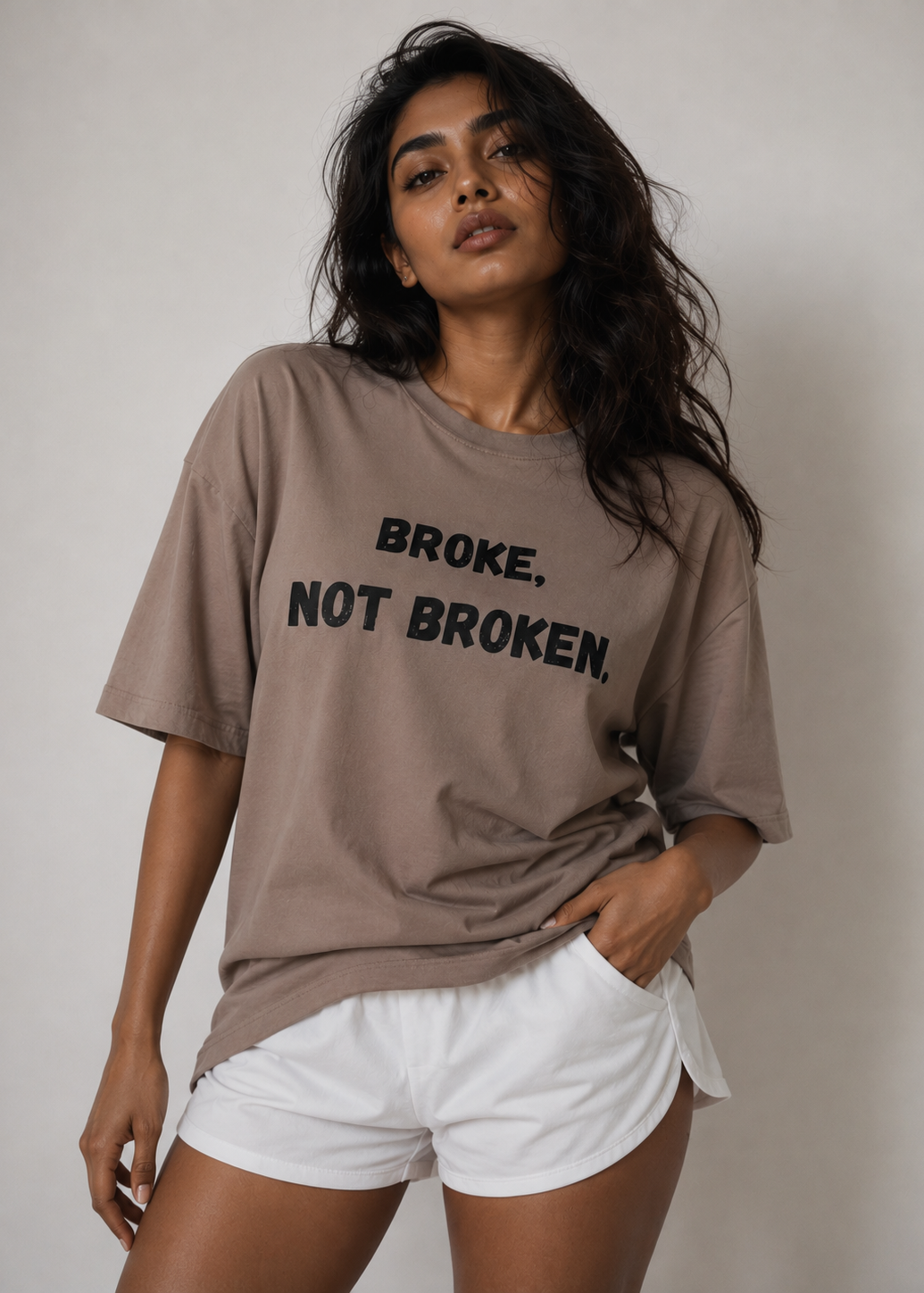 A woman wearing a taupe-brown oversized graphic t-shirt with the text 'BROKE. NOT BROKEN.