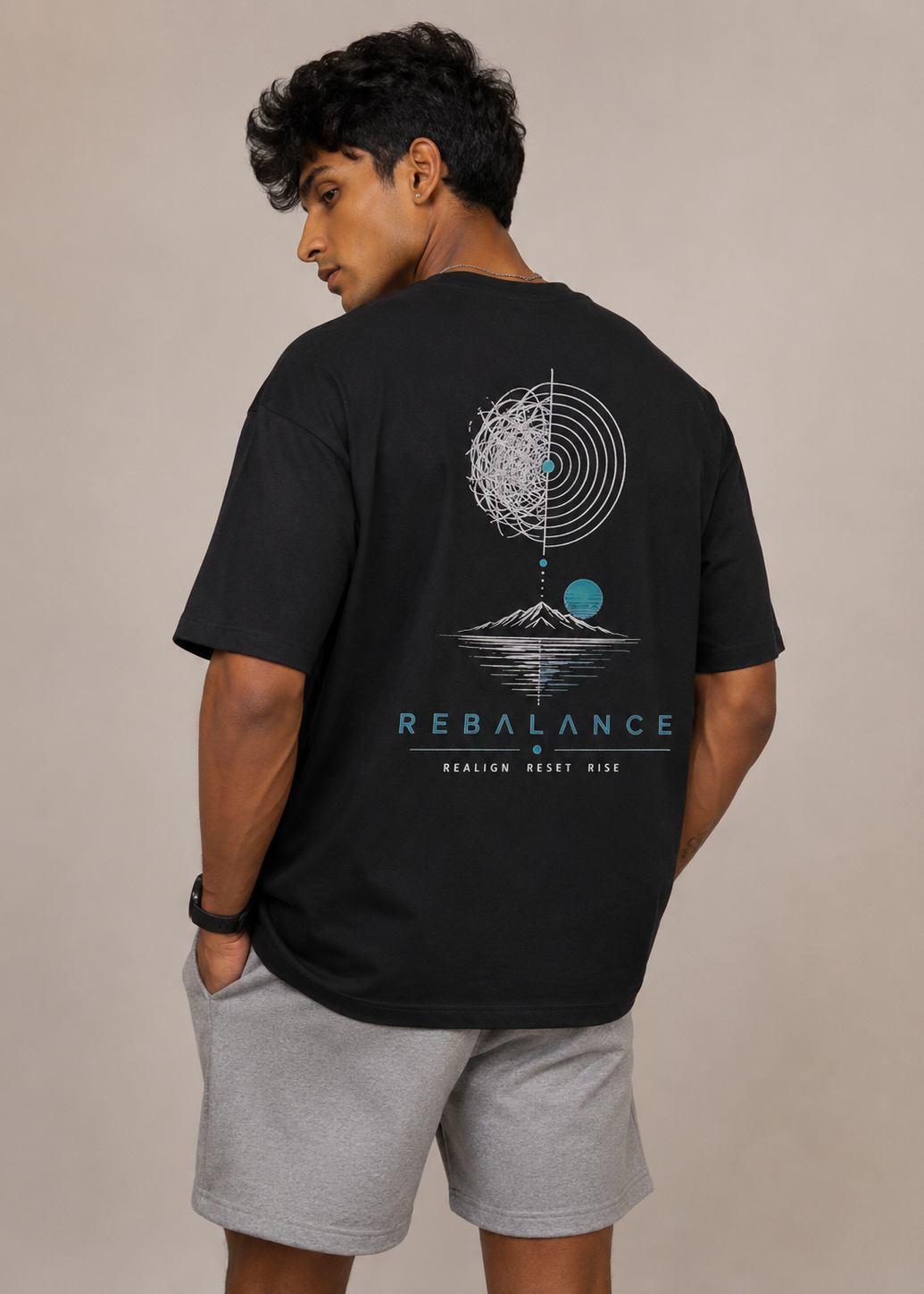 A man wearing a black t-shirt with the word 'REBALANCE' printed in light blue across the chest, paired with light gray heather shorts, standing against a neutral beige wall.