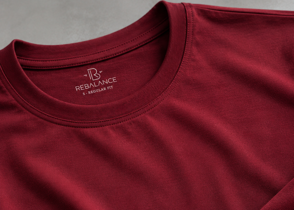A close-up of a burgundy red crewneck t-shirt by Bernalance, showing the inner neck label and a folded sleeve on a neutral gray background.