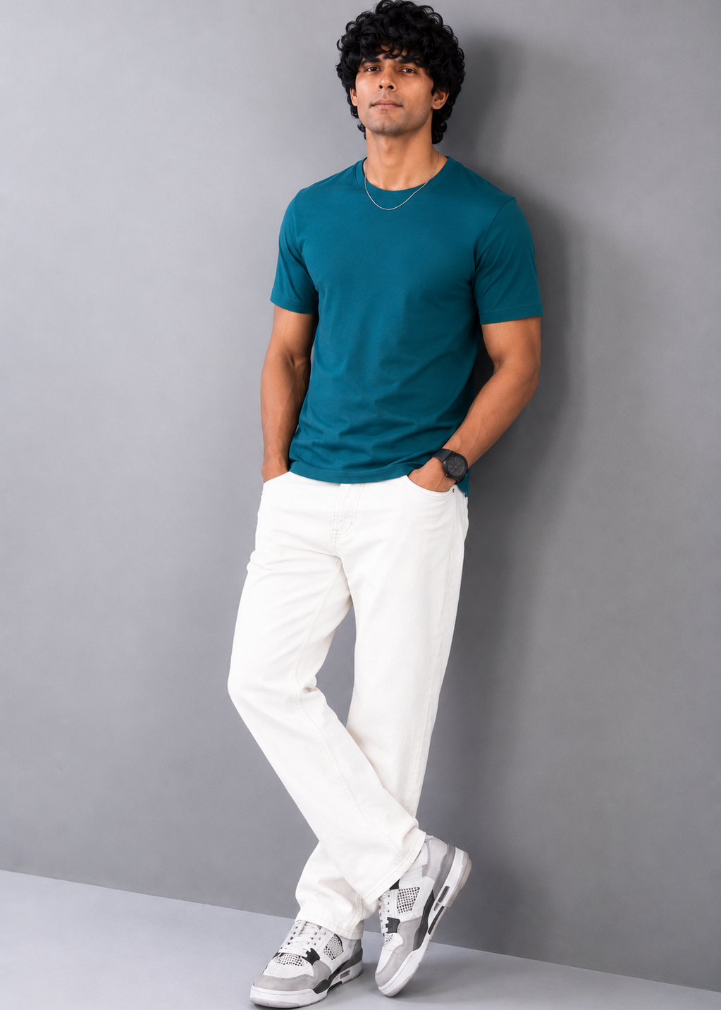 A man wearing a teal short-sleeve t-shirt, white trousers, and gray and white sneakers, standing against a plain gray wall.