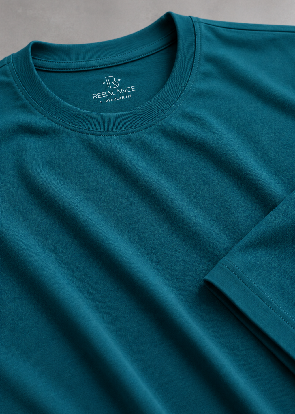 A close-up of a teal crew-neck t-shirt by REBALANCE, showing the inner neck label and a folded sleeve on a textured gray background.