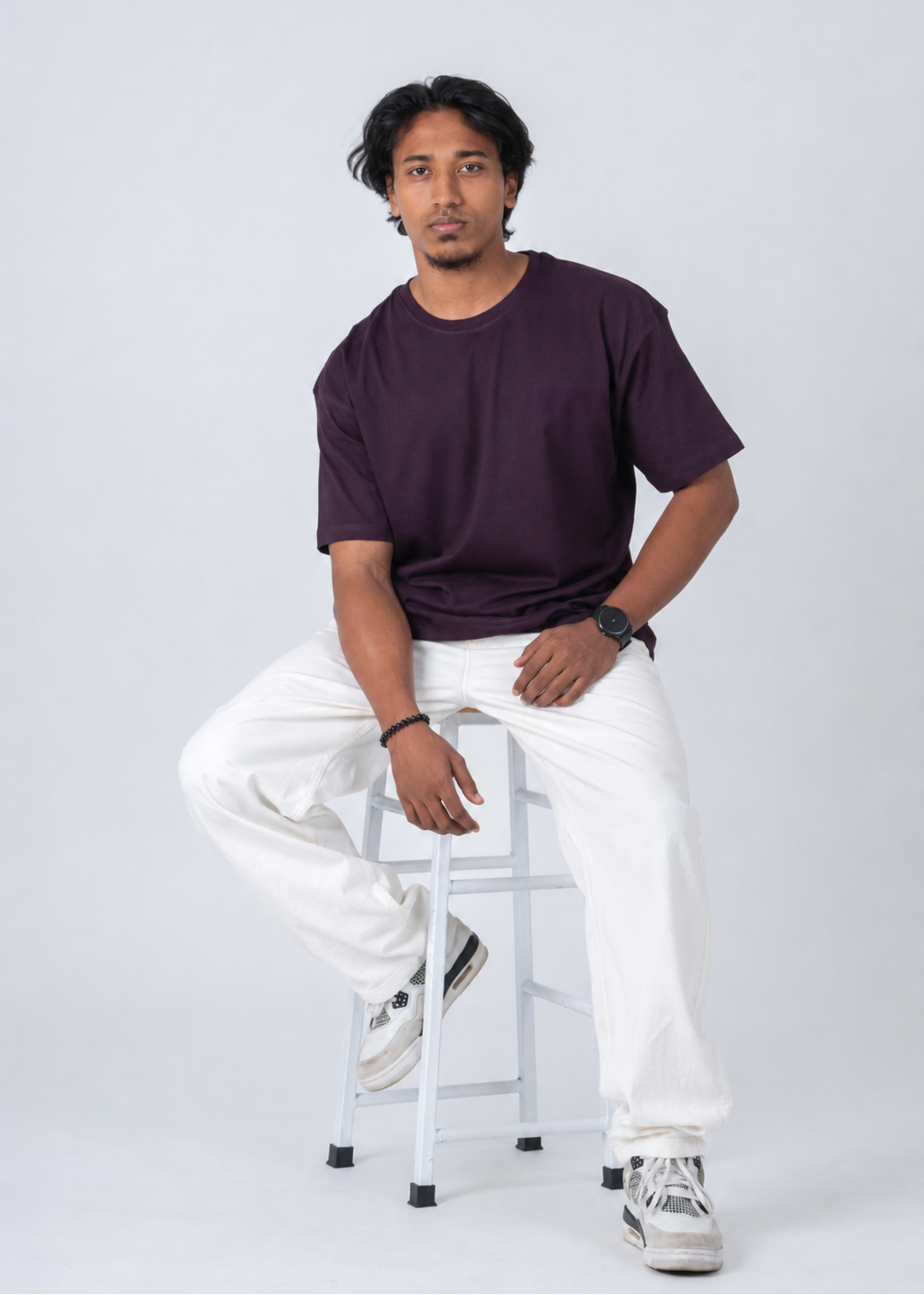 A man wearing a dark purple crewneck t-shirt, white sweatpants, and white sneakers sits on a white metal stool against a light gray studio background.