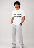 A man modeling a white crewneck t-shirt with black text that reads 'ZERO STRESS ZERO SAVINGS', paired with light gray sweatpants and gray sneakers, against a light gray studio background.
