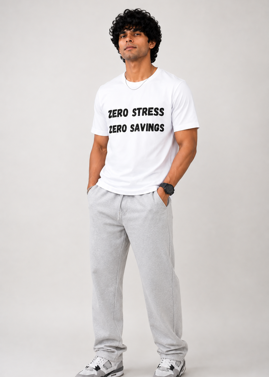 A man modeling a white crewneck t-shirt with black text that reads 'ZERO STRESS ZERO SAVINGS', paired with light gray sweatpants and gray sneakers, against a light gray studio background.