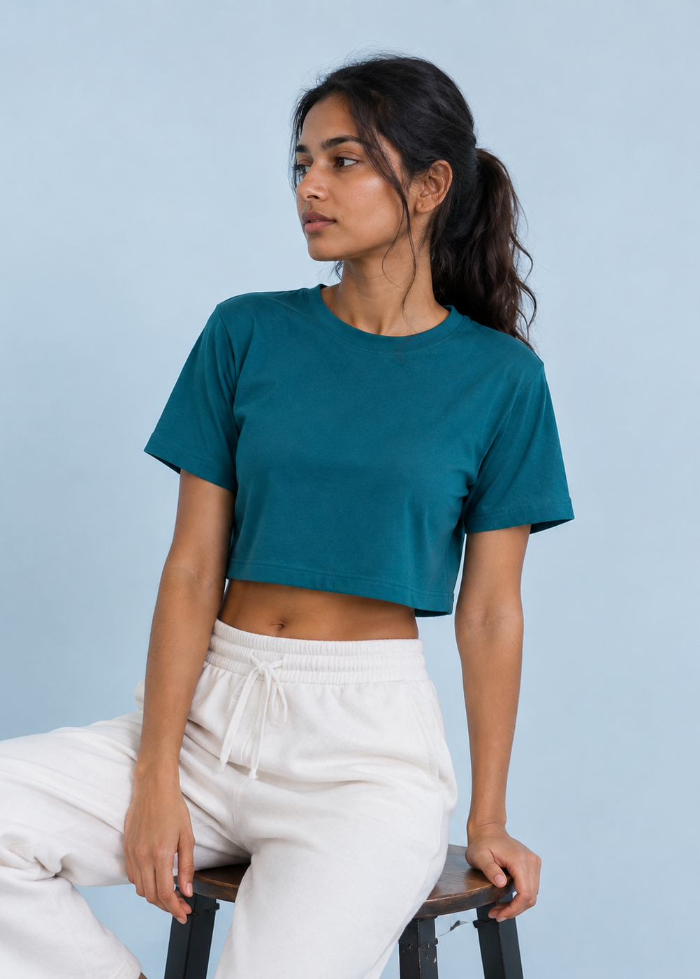 A woman wearing a teal short-sleeve crop top and white sweatpants, sitting on a stool against a light blue background.