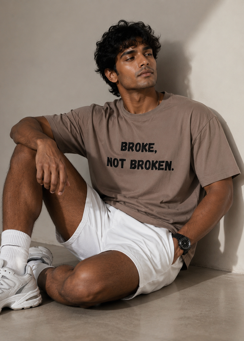 A man sitting on a concrete floor against a neutral wall, wearing a taupe brown graphic t-shirt with the text 'BROKE, NOT BROKEN.' in black, white shorts, white socks, and white New Balance sneakers.