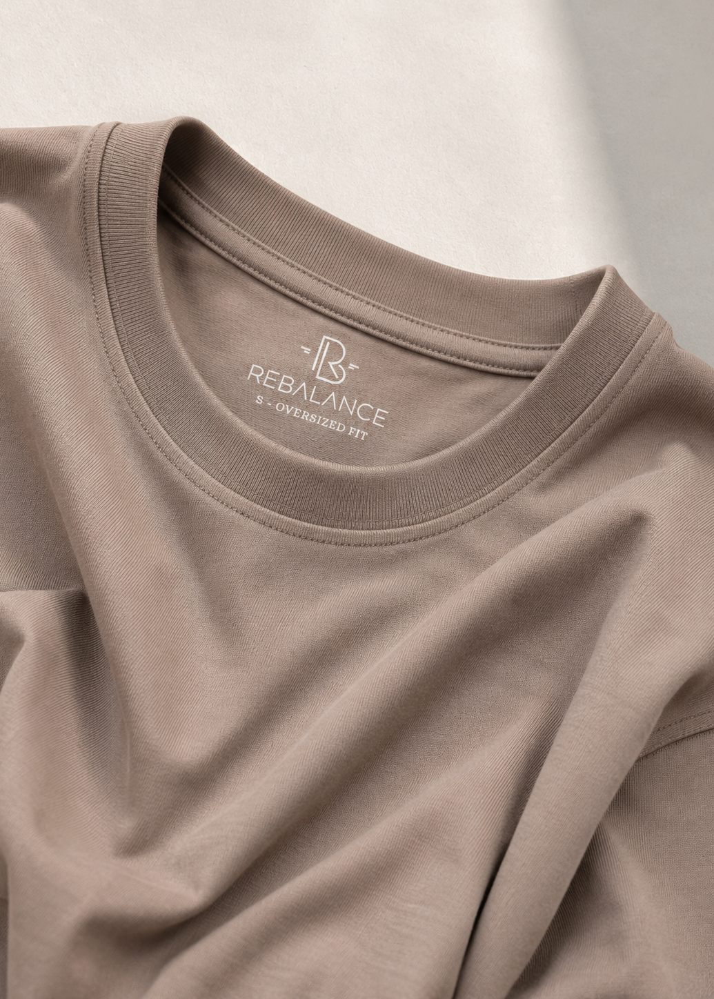 A close-up of a taupe crewneck t-shirt by REBALANCE, showing the inner neck label and soft fabric folds under warm, diffused light.