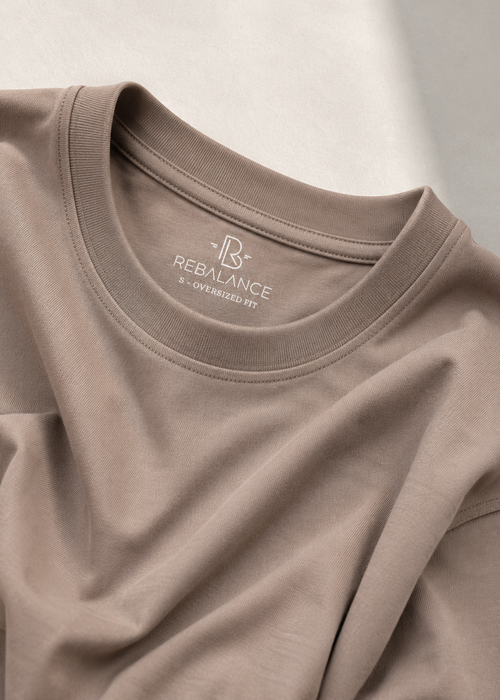 A close-up of a taupe crewneck t-shirt by REBALANCE, showing the inner neck label and soft fabric folds under warm, diffused light.