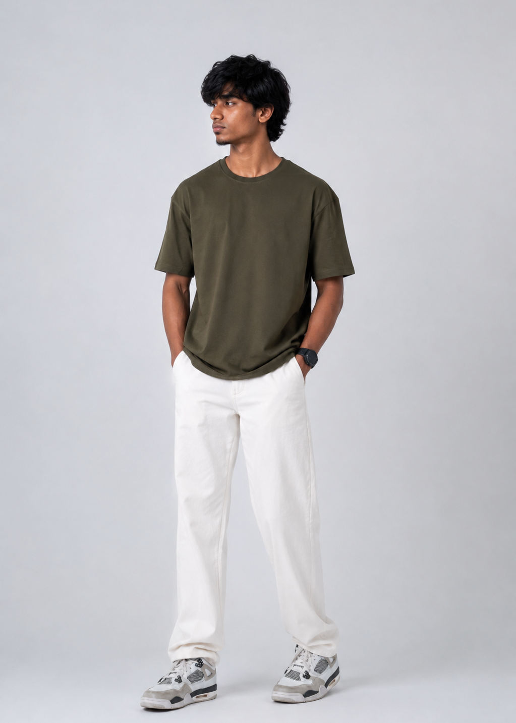 A man modeling a short-sleeve crewneck t-shirt in olive green, paired with off-white trousers and gray sneakers, standing against a light gray studio background.